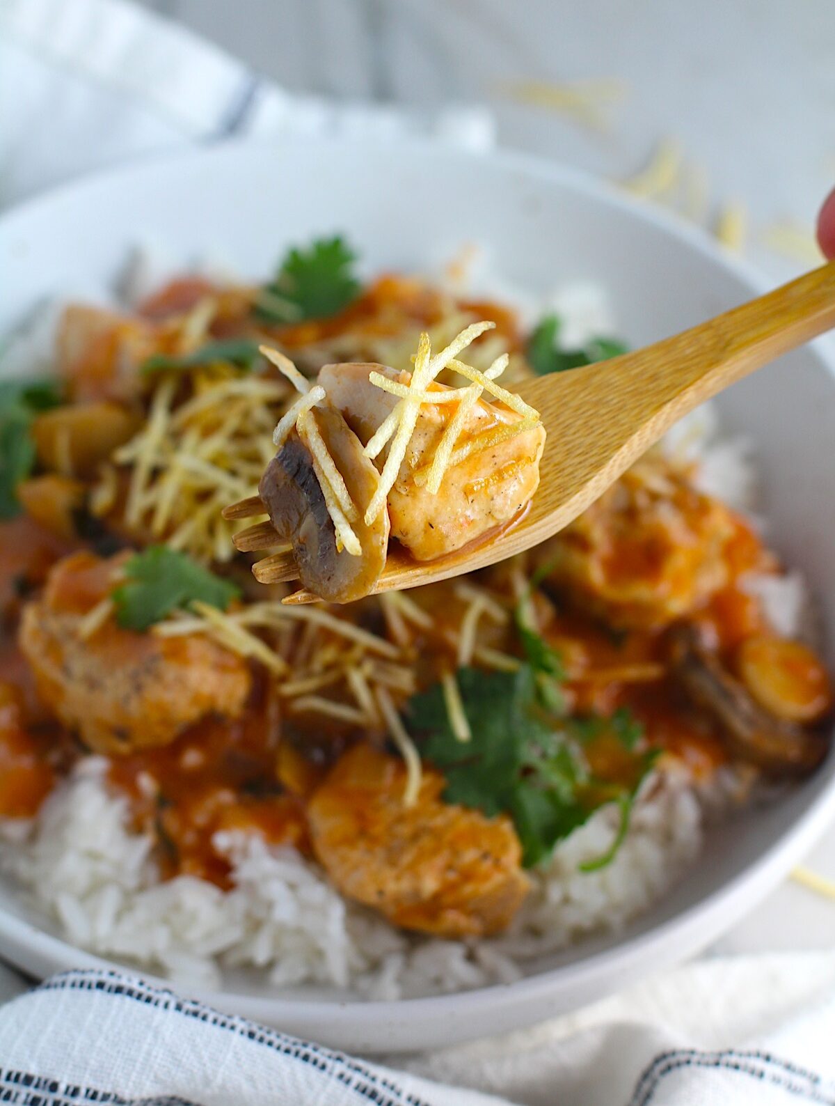 Fork holding bite of Brazilian Chicken Stroganoff in a tomato-based sauce over a bowl with white rice with cilantro garnish and thin potato sticks on top. This is an easy 30 minute dinner with a tomato based sauce, cream, and mushrooms all over rice.