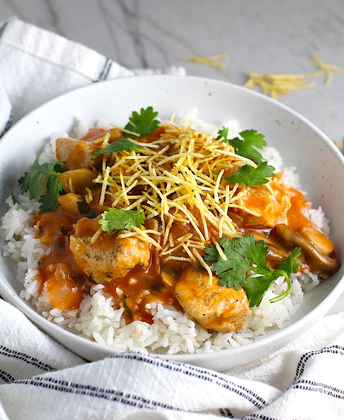 Brazilian Chicken Stroganoff in a tomato-based sauce in a bowl over white rice with cilantro garnish and thin potato sticks on top. This is an easy 30 minute dinner with a tomato based sauce, cream, and mushrooms all over rice.