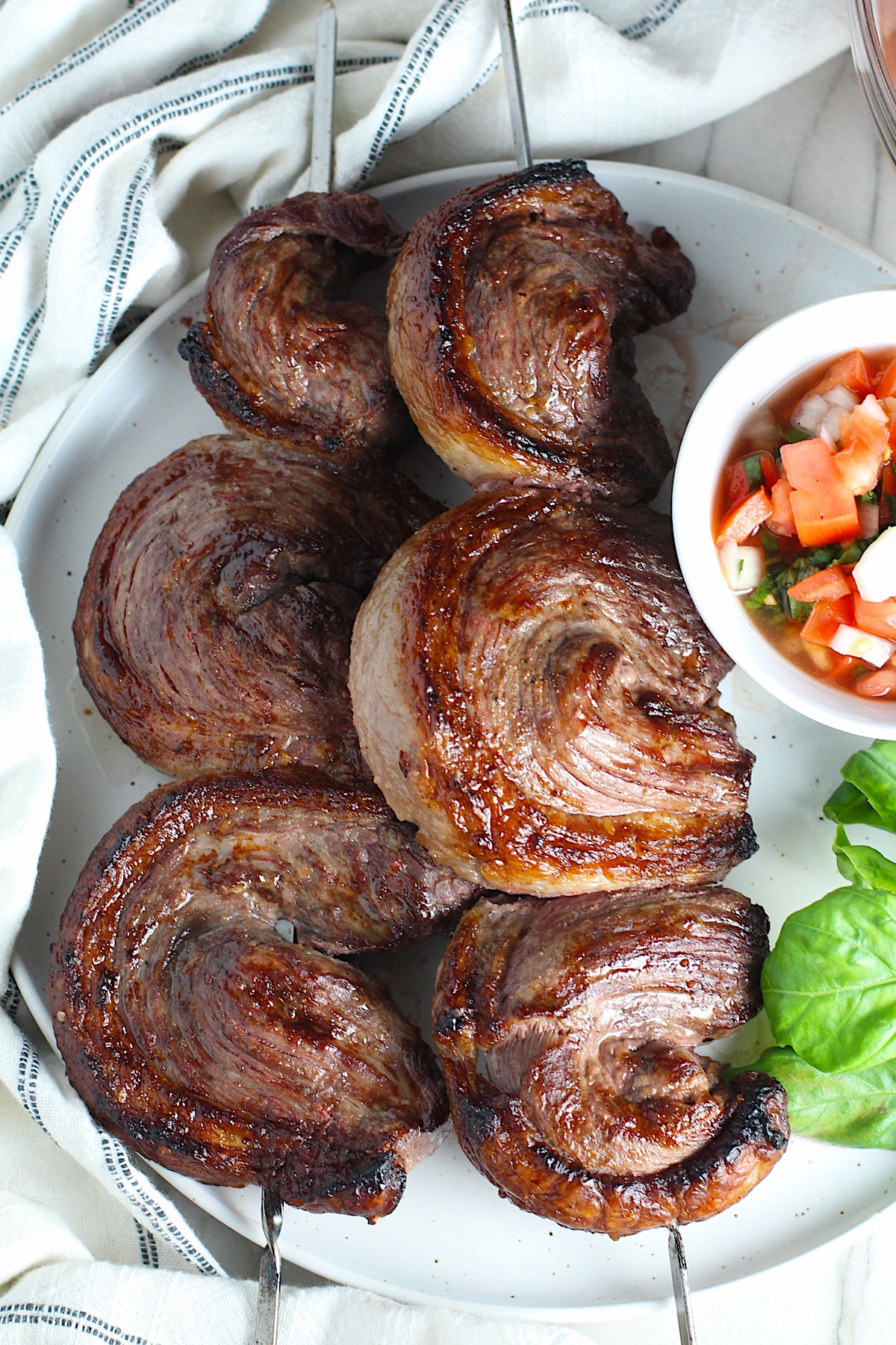 Picanha steak skewered on a plate with a tomato salad and basil leaves.  It's the best Brazilian steak recipe, a tender and delicious cut of beef seasoned with a Brazilian blend and grilled perfectly!