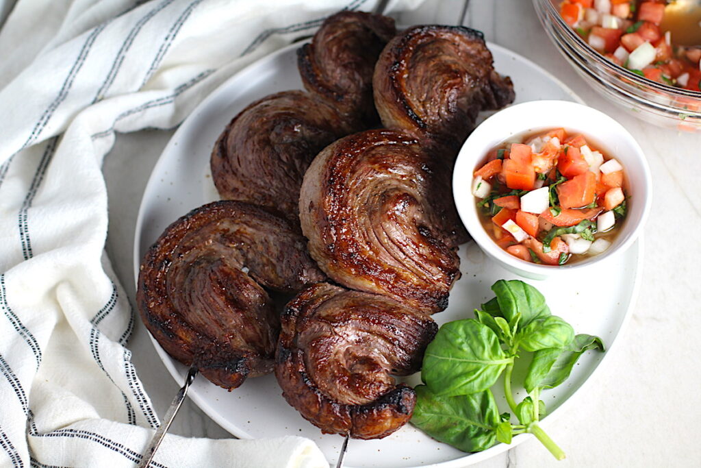 Picanha steak skewered on a plate with a tomato salad and basil leaves. It's the best Brazilian steak recipe, a tender and delicious cut of beef seasoned with a Brazilian blend and grilled perfectly!