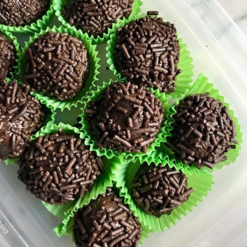 Brigadeiros in green paper in a plastic container. This Brigadeiro Recipe is based on traditional Brazilian chocolate truffles rolled in sprinkles. You won't believe how easy they are to make!