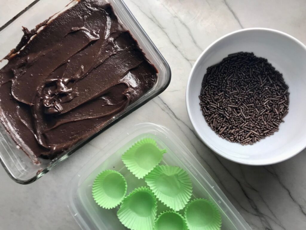 Dish with brigadeiro spread out with bowl of sprinkles next to it, and container with green paper cups. This Brigadeiro Recipe is based on traditional Brazilian chocolate truffles rolled in sprinkles. You won't believe how easy they are to make!