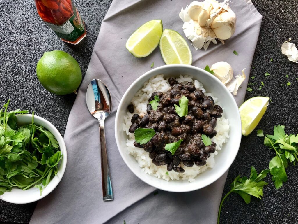 Brazilian Black Beans Recipe (Feijao) over rice in a bowl with cilantro leaves on top, lime wedges and garlic cloves on the side, and a hot sauce bottle. It's delicious, thick, & hearty.