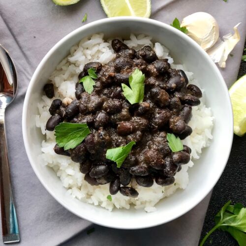 Brazilian Black Beans Recipe (Feijao) over rice in a bowl with cilantro leaves on top, lime wedges and garlic cloves on the side. It's delicious, thick, & hearty.