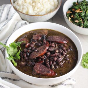 Bowl of cooked black beans with pork sausage slices and beef and cilantro garnish on side. It is on table with a bowl of cooked kale and a bowl of rice with towel next to it. Feijoada is rice and beans Brazilian style with pork sausages, beef, and more.