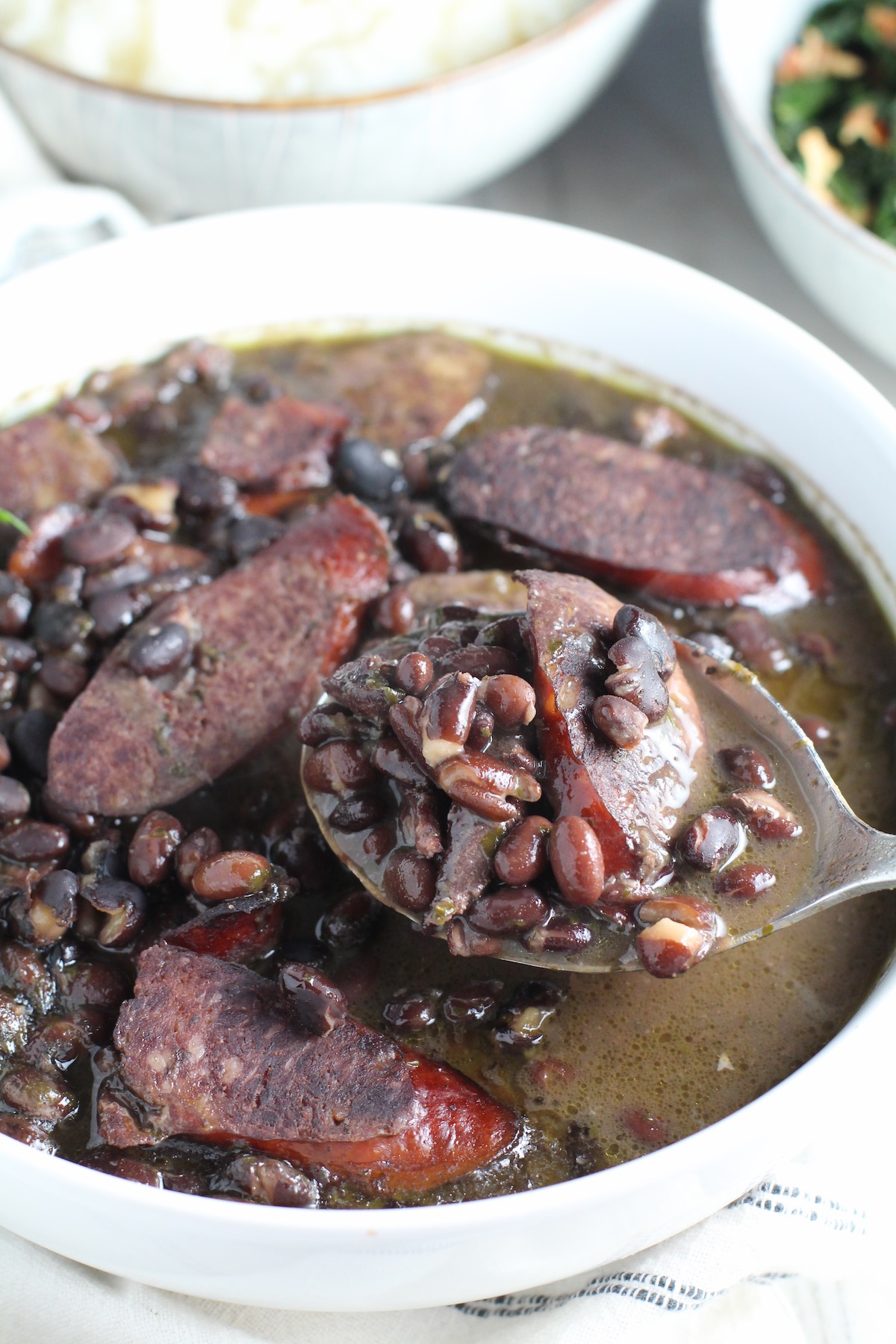 Serving spoon scooping up cooked black beans with pork sausage and beef in a white bowl. Feijoada is rice and beans Brazilian style with pork sausages, beef, and more.