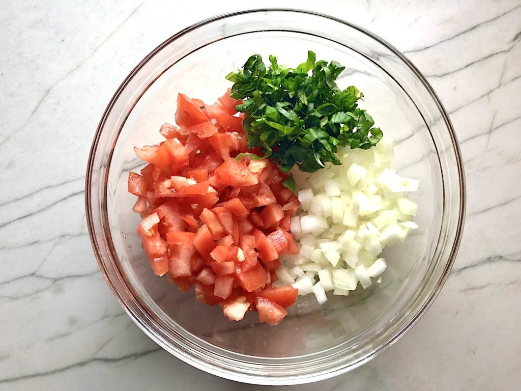 Diced tomato, diced onion, basil in sections in a clear bowl for Tomato and onion salad in clear bowl on counter with towel.
