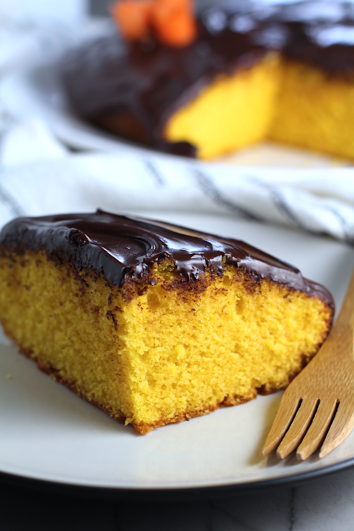 Slice of Brazilian Carrot Cake with Chocolate Sauce topping on a plate with bamboo fork and whole cake in background. It's absolutely delicious and easy to make!