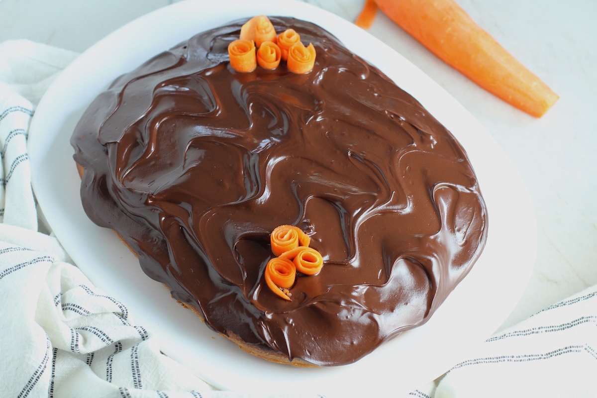 Whole Brazilian Carrot Cake with Chocolate Sauce topping and carrot rose decorations on top on a platter on counter with peeled carrot next to it. It's absolutely delicious and easy to make!