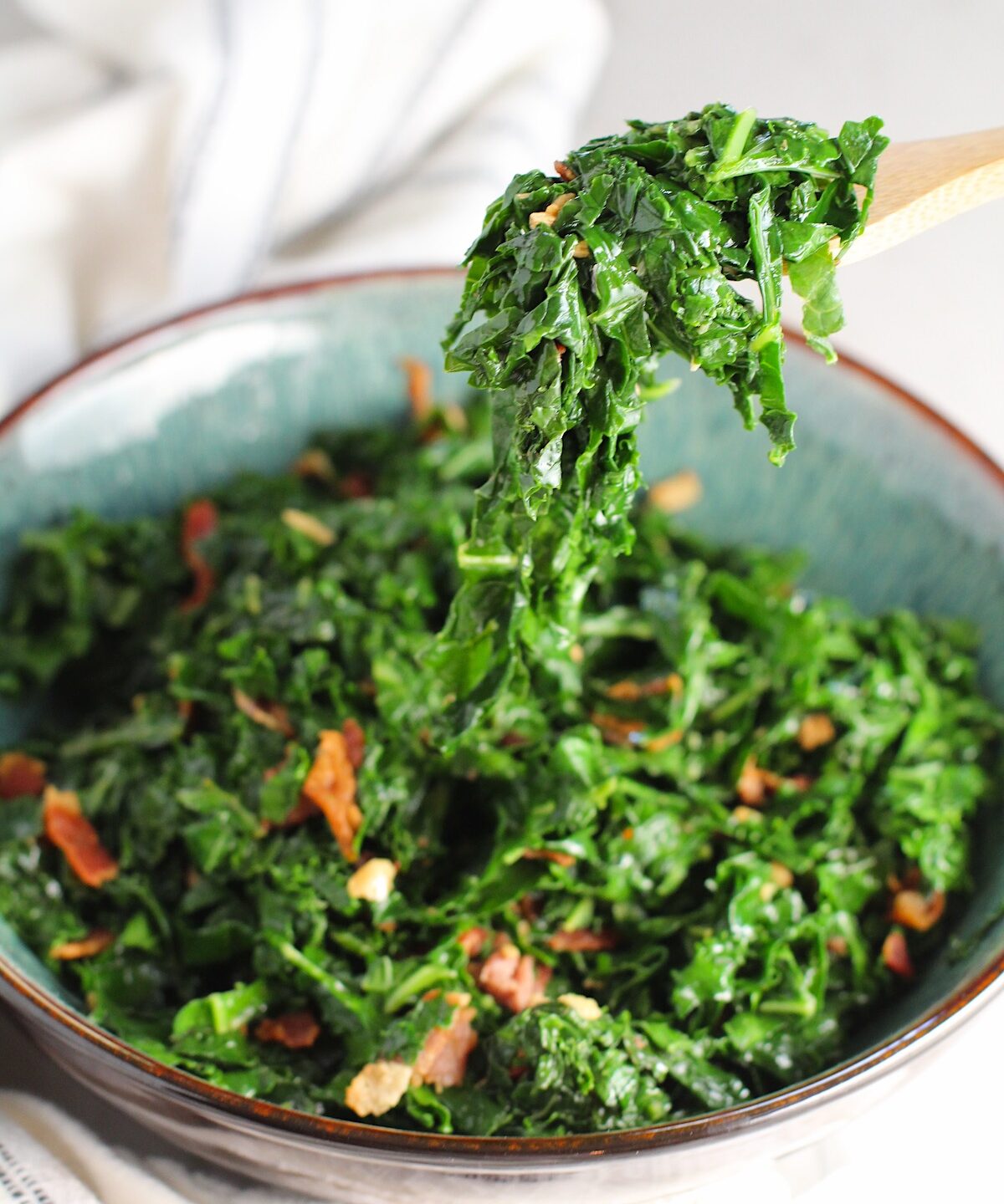 Spoon scooping Brazilian sauteed Kale with Bacon and Garlic in a bowl on counter with white and blue towel. It's known as Couve Mineira in Portuguese, and is an easy and delicious side for any dinner!