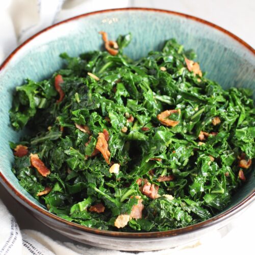 Brazilian sauteed Kale with Bacon and Garlic in a bowl on counter with white and blue towel. It's known as Couve Mineira in Portuguese, and is an easy and delicious side for any dinner!