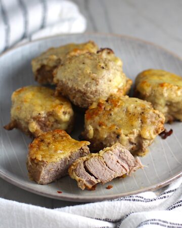 Parmesan Pork Tenderloin pieces on a plate with one cut in half. They are juicy, salty, and slightly cheesy. It's easy to make in the oven so you can enjoy it at home anytime!