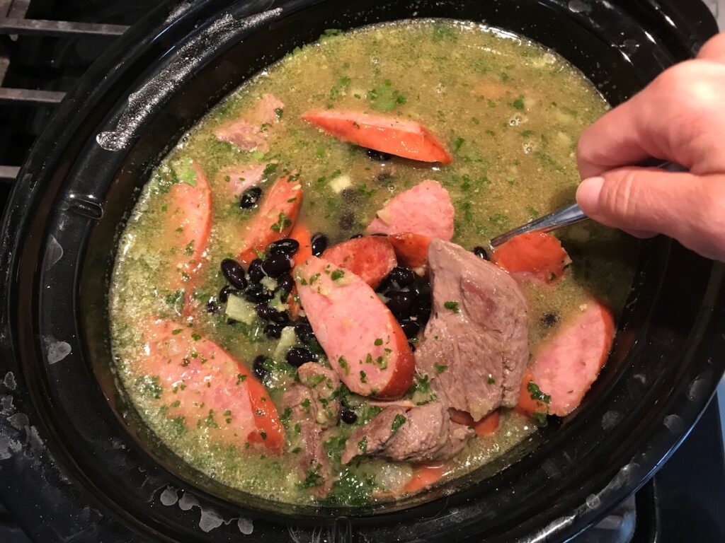 Hand using spoon to mix broth, beans, beef cubes and pork sausage, and other ingredients in slow cooker for Feijoada recipe. It is rice and beans Brazilian style with pork sausages, beef, and more.
