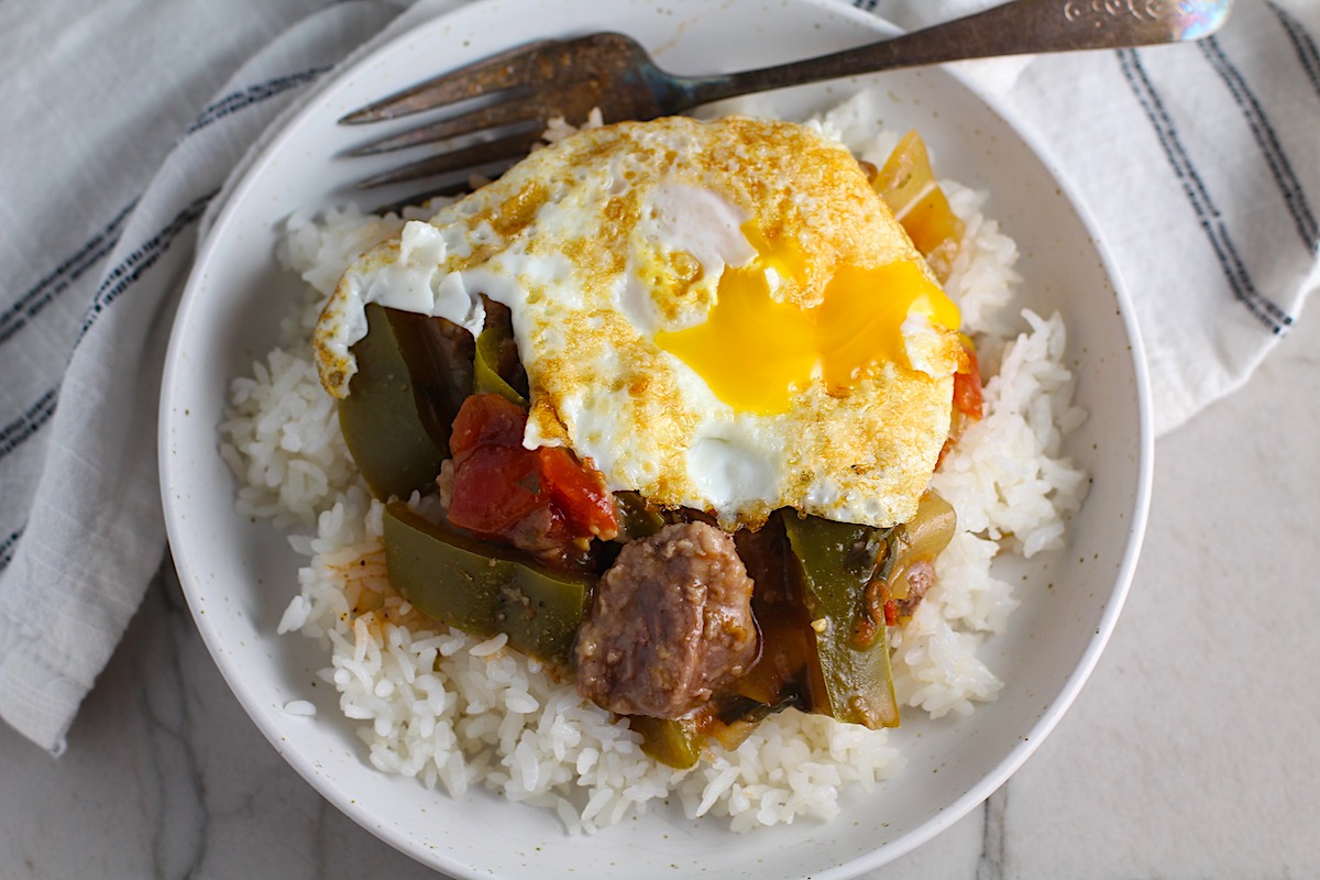 Fried egg on top of cooked Brazilian Beef Stew Stove Top recipe over rice in bowl with fork in it on counter. It's a savory, succulent and hearty dish that you can make anytime!