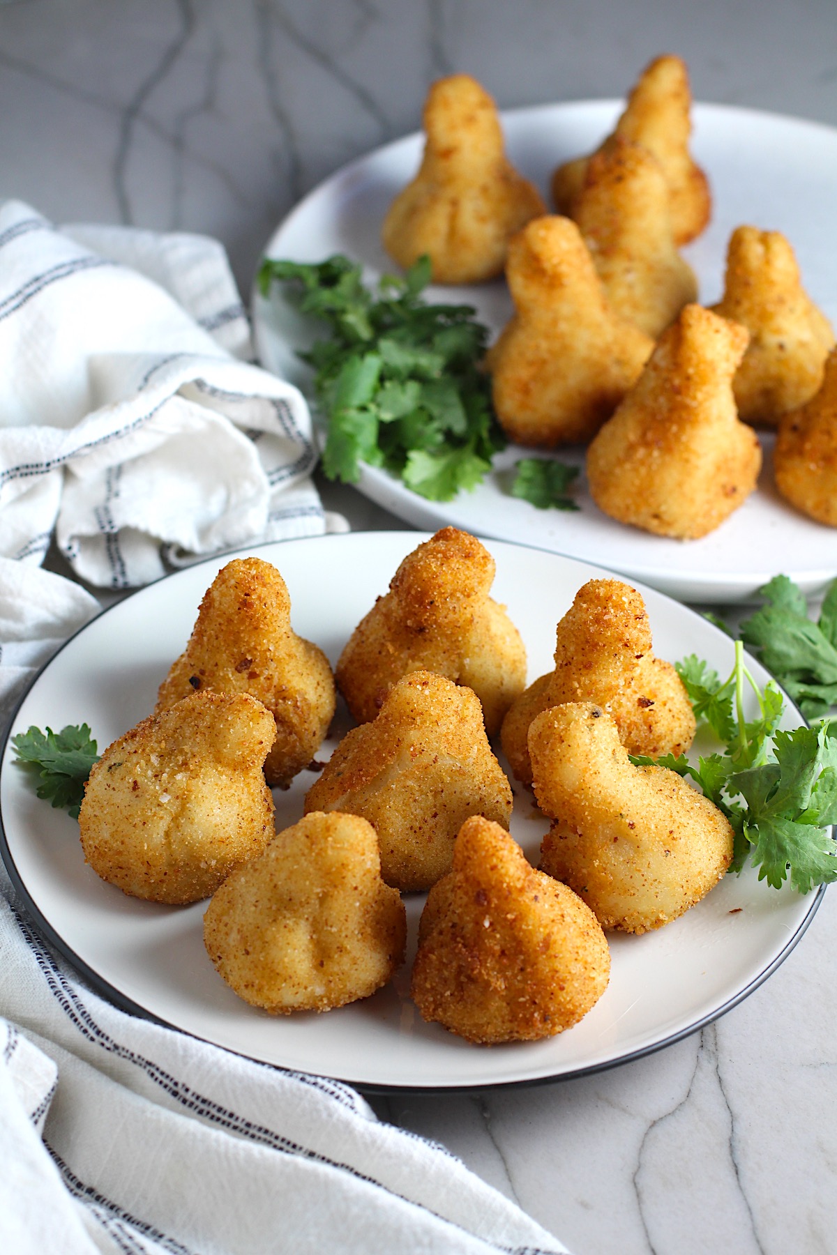 Golden brown breaded Brazilian Coxinha Chicken Croquettes on two plates with cilantro garnish on a counter with towel next to it. 
