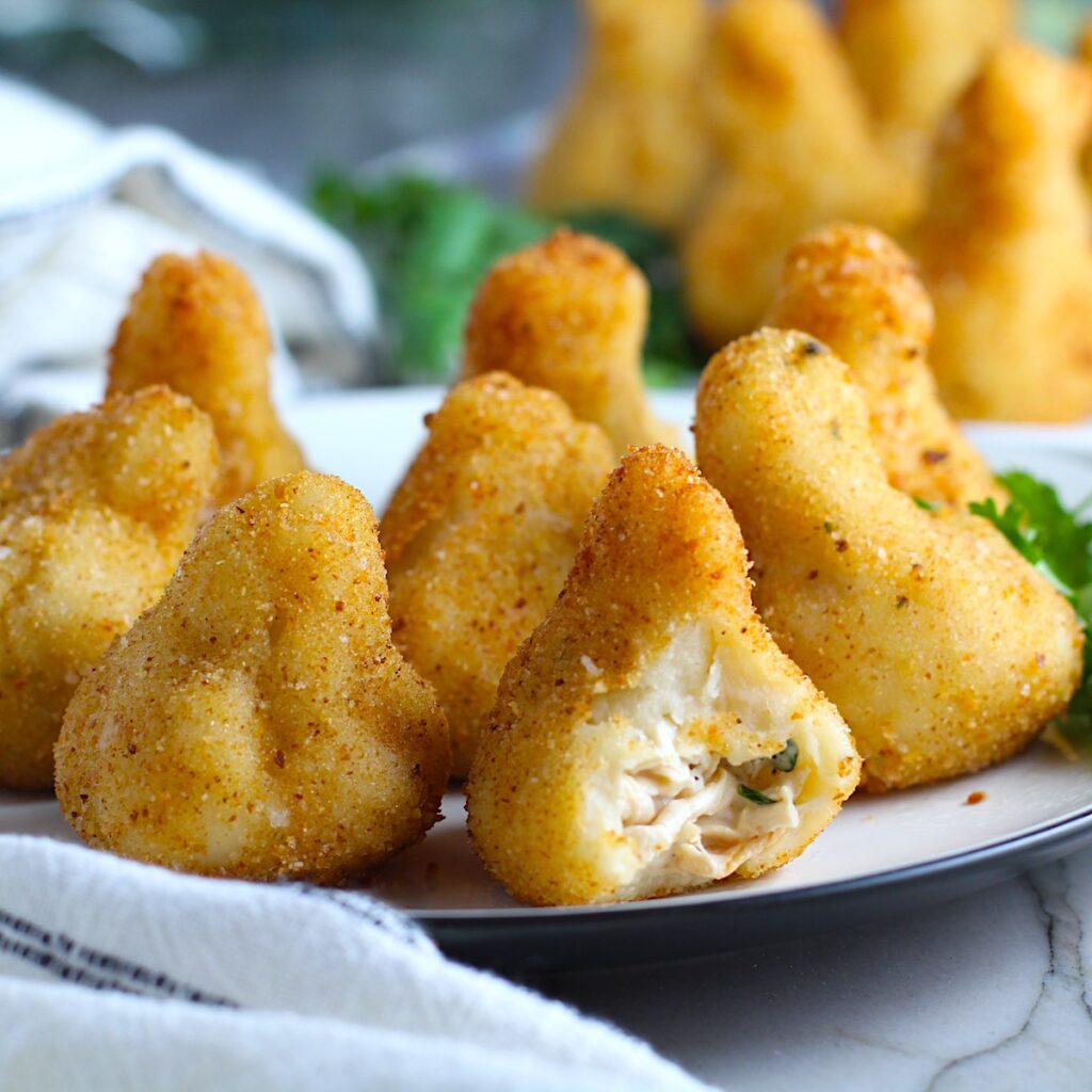 Golden brown breaded Brazilian Coxinha Chicken Croquettes on a plate with a bite taken out of the croquette in front. The plate has cilantro garnish and it's on counter with towel next to it.