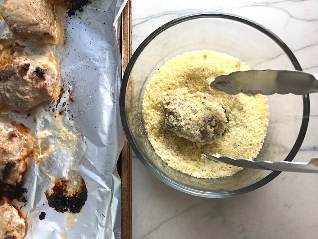 Cooked Pork tenderloin pieces on a pan with one piece being coated in a bowl of grated parmesan cheese next to it for Parmesan Pork Tenderloin recipe.