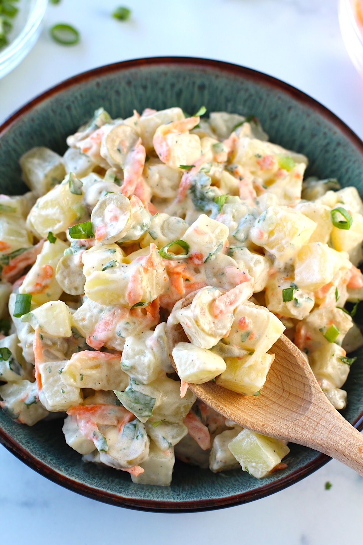 Brazilian Potato Salad in a bowl with carrots, green olives, and a mayonnaise dressing, with bamboo spoon scooping some up and cilantro garnish on top.