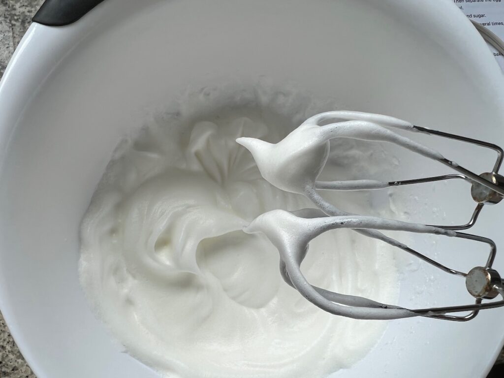 Egg whites beaten to a stiff peak on the mixer whisks above the whites in bowl for Brazilian Coconut Cake.