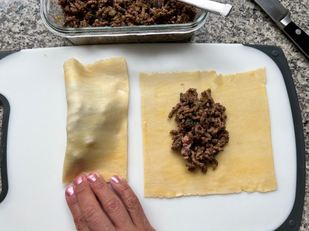 Fingers pressing the edges of the folded pastel dough with a second pastel laying open next to it with filling on top for Brazilian Pastel recipe.