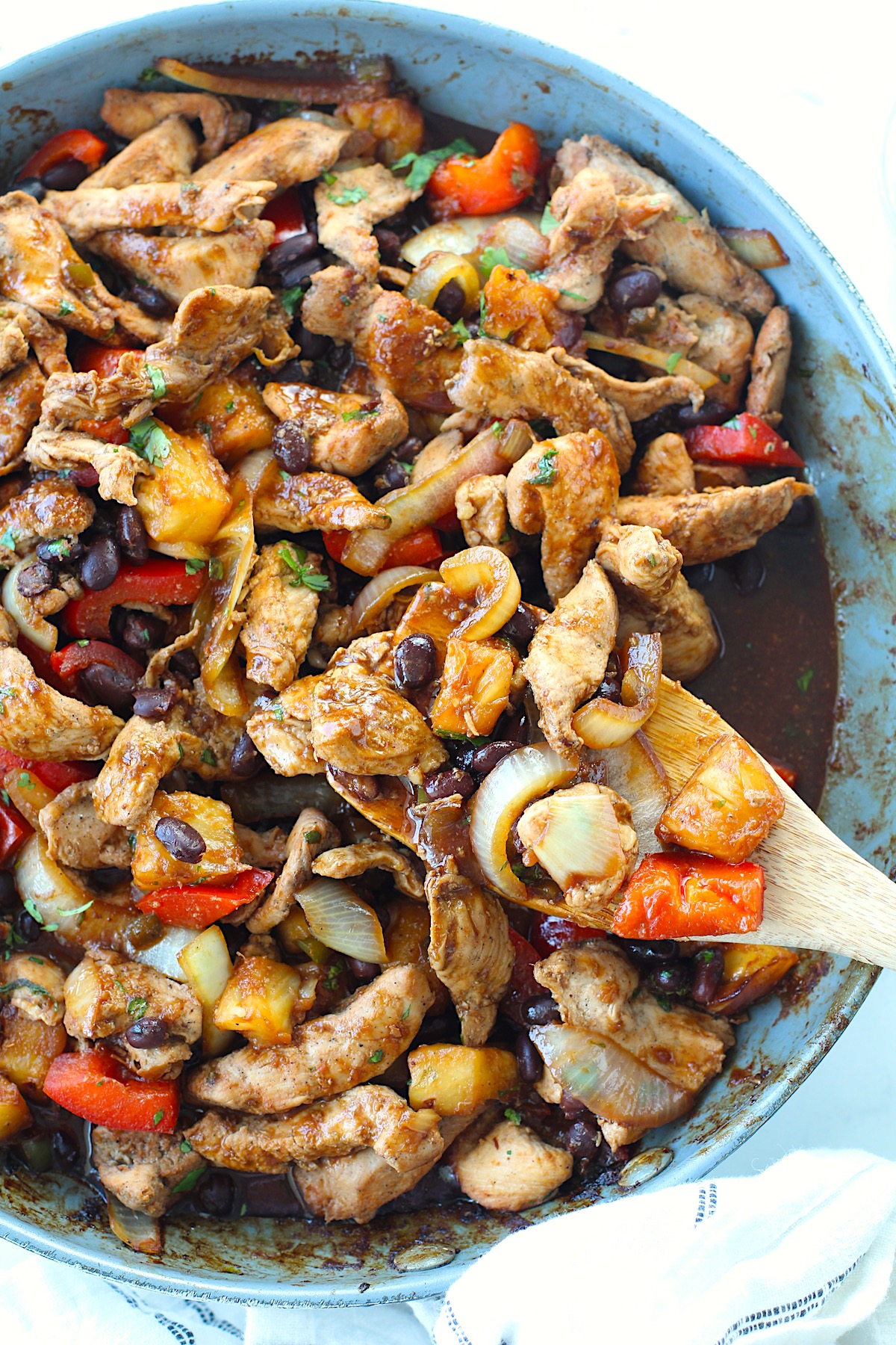 Brazilian Marinated Chicken Stir Fry with pineapple, black beans, red Pepper and onions in a skillet with spatula with cilantro garnish. It is truly one amazing dish with incredible flavor!