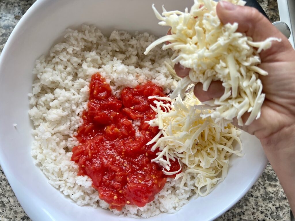 Hand adding shredded mozzarella to crushed tomatoes and cooked rice in a bowl for Baked Rice with Cheese and Tomatoes.