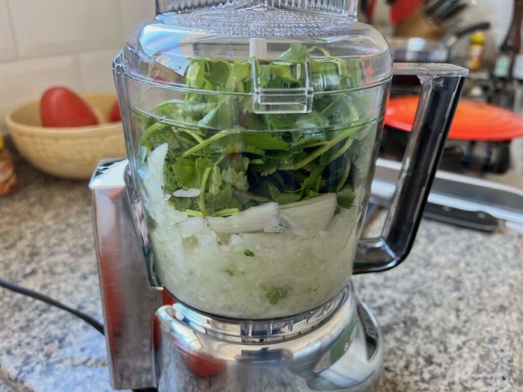 Side view of food processor with onions on the bottom, garlic, and cilantro leaves on top for Carioca Beans recipe.