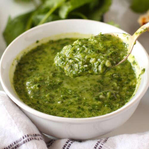 Spoon scooping Brazilian Cilantro Chimichurri recipe over bowl with fresh cilantro and bottle of olive oil in background.
