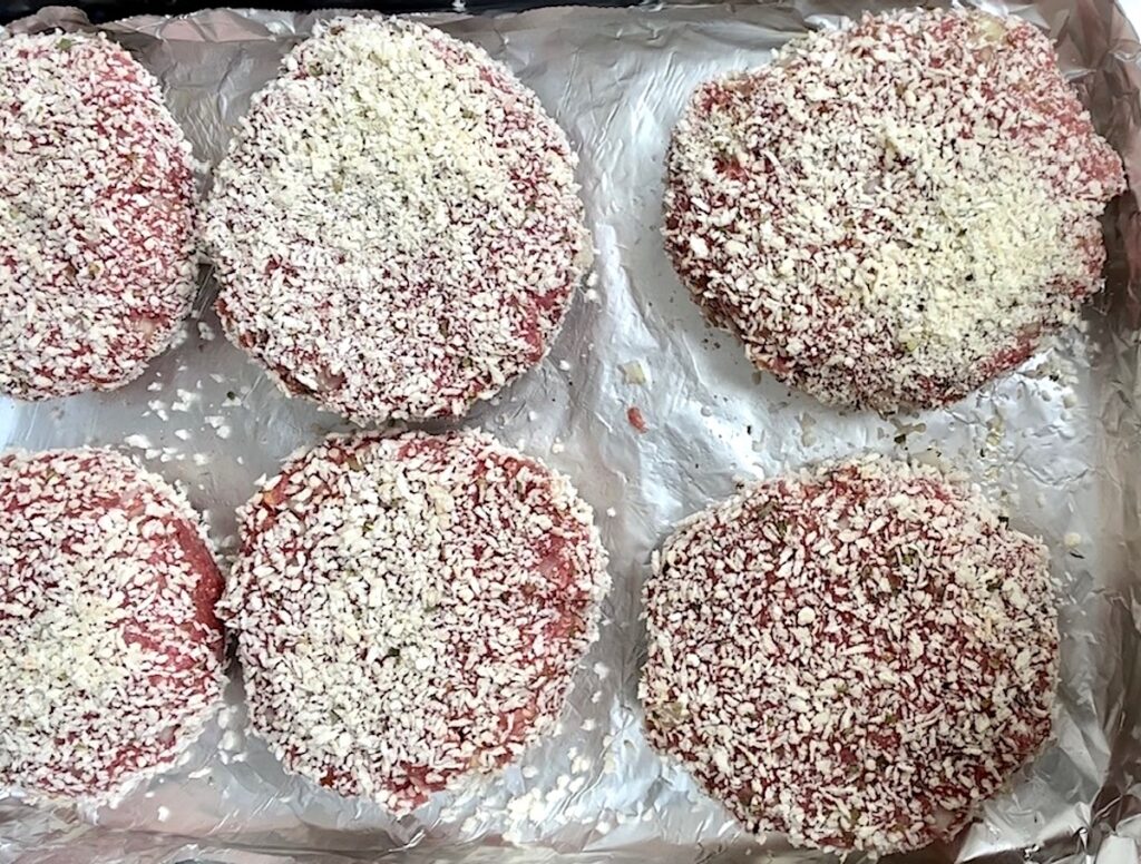 Six polpettone beef patties that have been coated in breadcrumbs laying on an aluminum lined sheet pan before cooking.