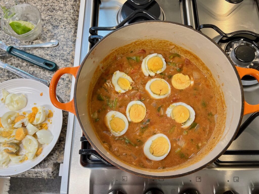 Sliced hard boiled eggs added to Moqueca de Ovos in a pot on stove with plate of slice eggs on counter next to it.