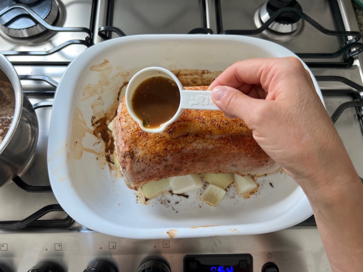 Hand holding a ¼ cup measuring cup with orange sauce about to pour over a seasoned raw pork ribeye roast on top of cut onions in a casserole dish.