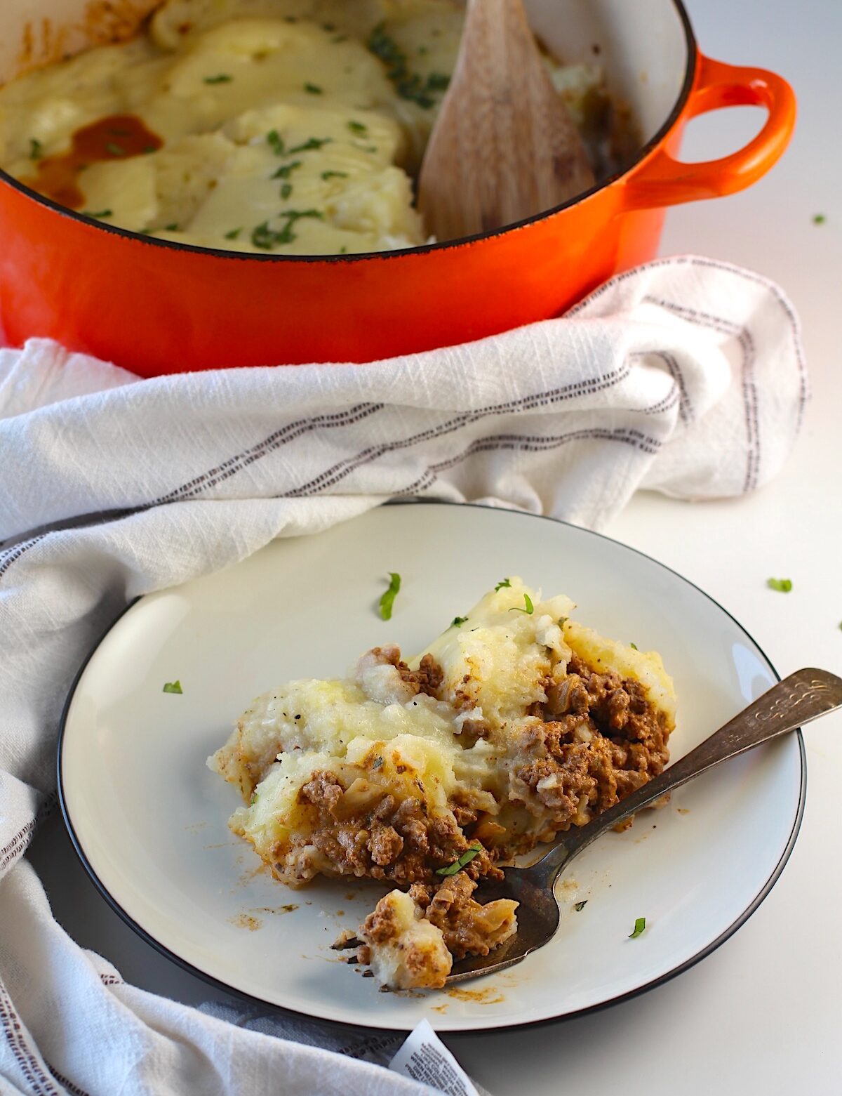Piece of Dutch oven shepherd's pie with ground beef filling topped with mashed potatoes on a plate with fork laying down with a bite. Orange coated dutch oven with the shepherd's pie is in the background.