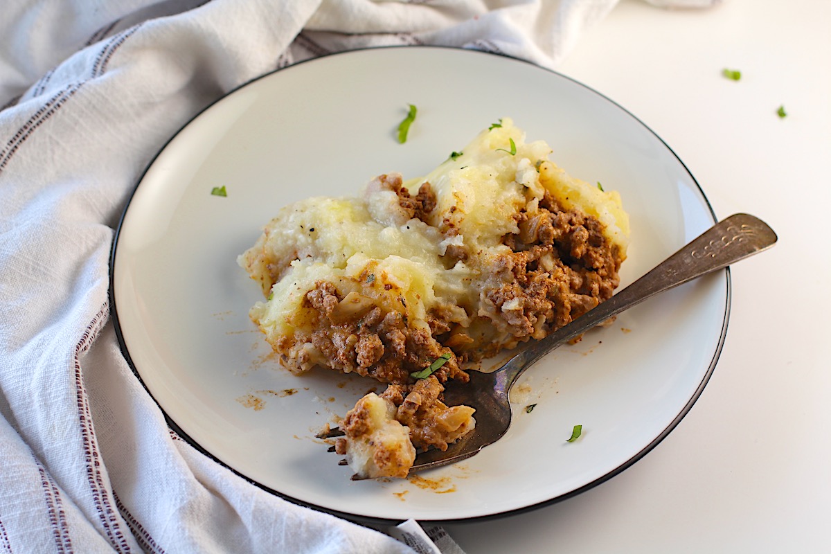 Piece of Dutch oven shepherd's pie with ground beef filling topped with mashed potatoes on a plate with fork laying down with a bite.