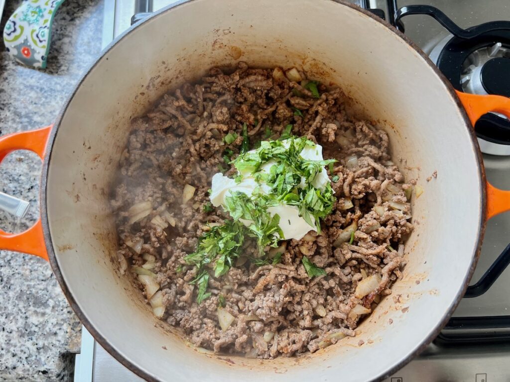 Ground beef cooked with a spoonful of cream cheese on top and fresh cilantro in dutch oven on stove for Dutch oven Shepherd's Pie.