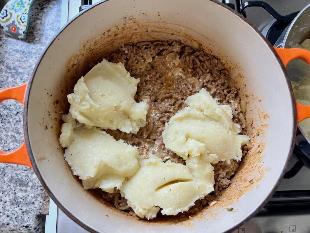 Scoops of mashed potatoes added on top of ground beef filling in a dutch oven on stove for Dutch oven Shepherd's Pie.