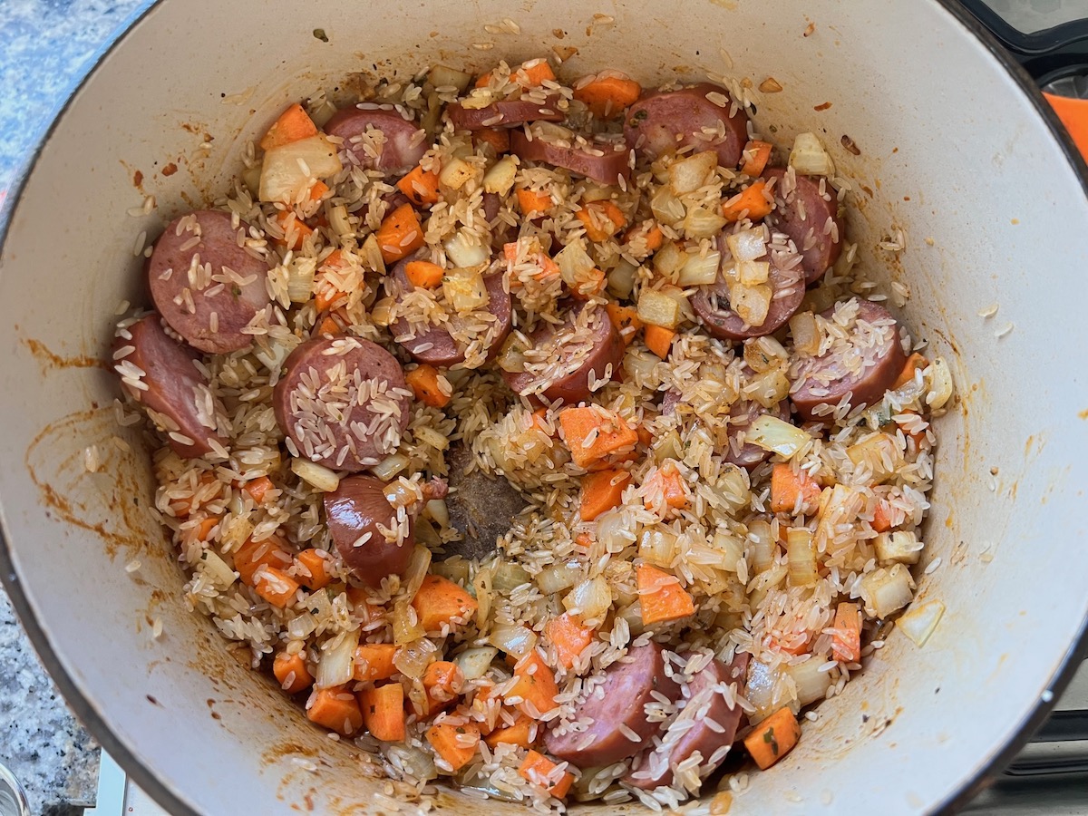 Uncooked rice mixed into sausage, carrots, onions, and garlic cooking in a pot for Galinhada, Dutch Oven Chicken and Rice.