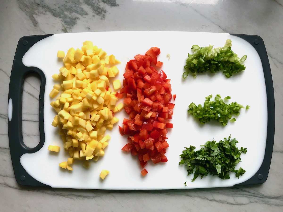 Mango, red pepper, cilantro, jalapeno, and scallions all chopped up and separated on a cutting board for Spicy Mango Salsa recipe