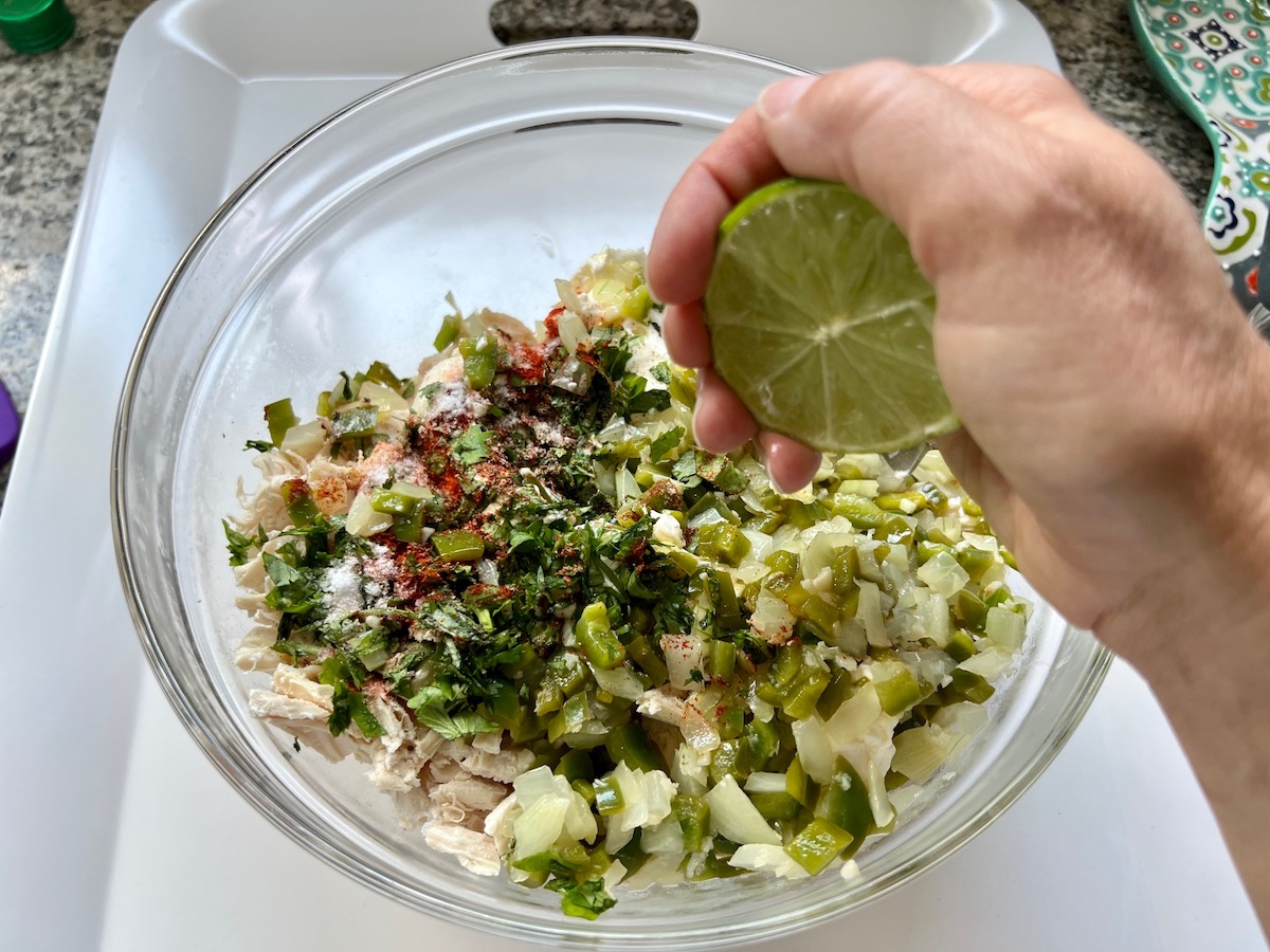Hand squeezing lime over a bow filled with shredded chicken, cream cheese, cilantro, onions, green pepper, and seasonings for the Ultimate Chicken Casserole Recipe.