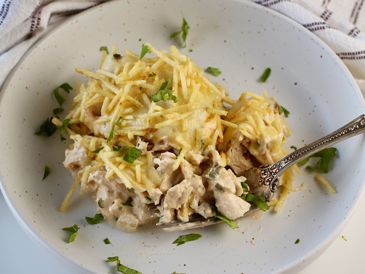 Ultimate Chicken Casserole recipe on a plate with a fork. It has shredded chicken, green peppers, onions, garlic, cilantro, paprika, and cream cheese holding it all together. On top, you get a layer of ooey gooey cheese and then crunchy potato sticks!