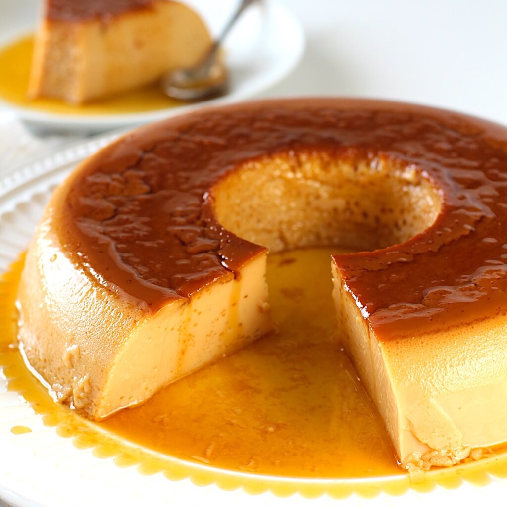 Brazilian Flan recipe in a tube shape with caramel sauce on top on a platter with a piece cut out of the front. In the background is the piece on a plate with a spoon.