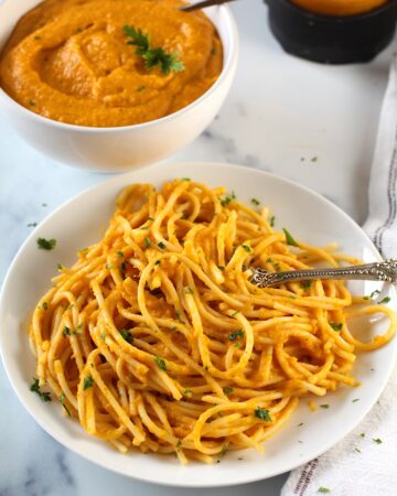 Plate of pasta mixed with carrot sauce with a fork in pasta and a napkin on counter. In the background is a bowl of carrot sauce with a spoon and parsley garnish. Also in background is the blender with the sauce in the bottom.