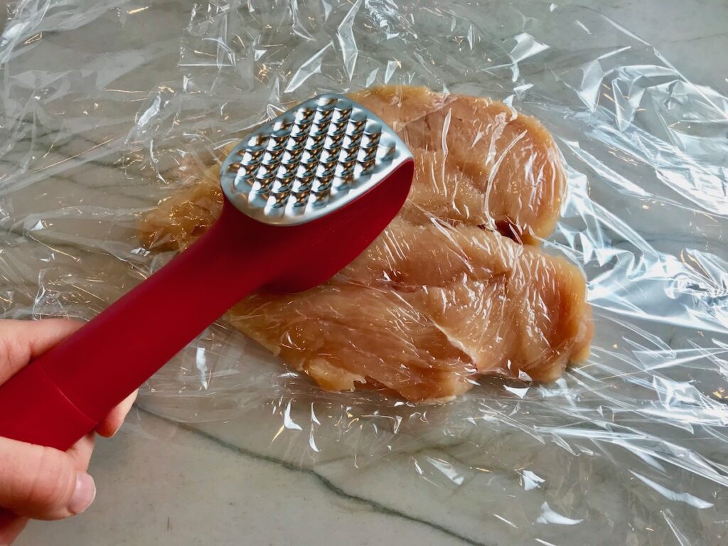 Butterflied chicken breast between two pieces of plastic wrap with red handled meat tenderizer pounding the outside of the chicken for Mozzarella Stuffed chicken.