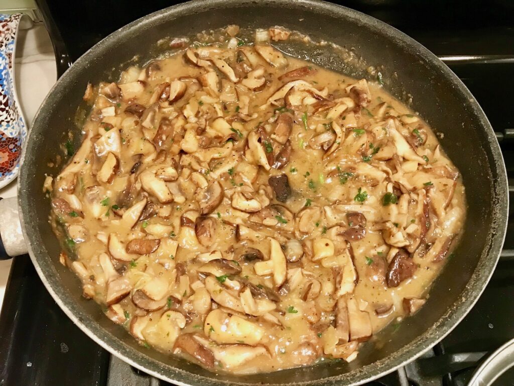 Sliced shiitake and cremini mushrooms cooking with sauce in a large skillet on the stove for Mozzarella Stuffed Chicken.