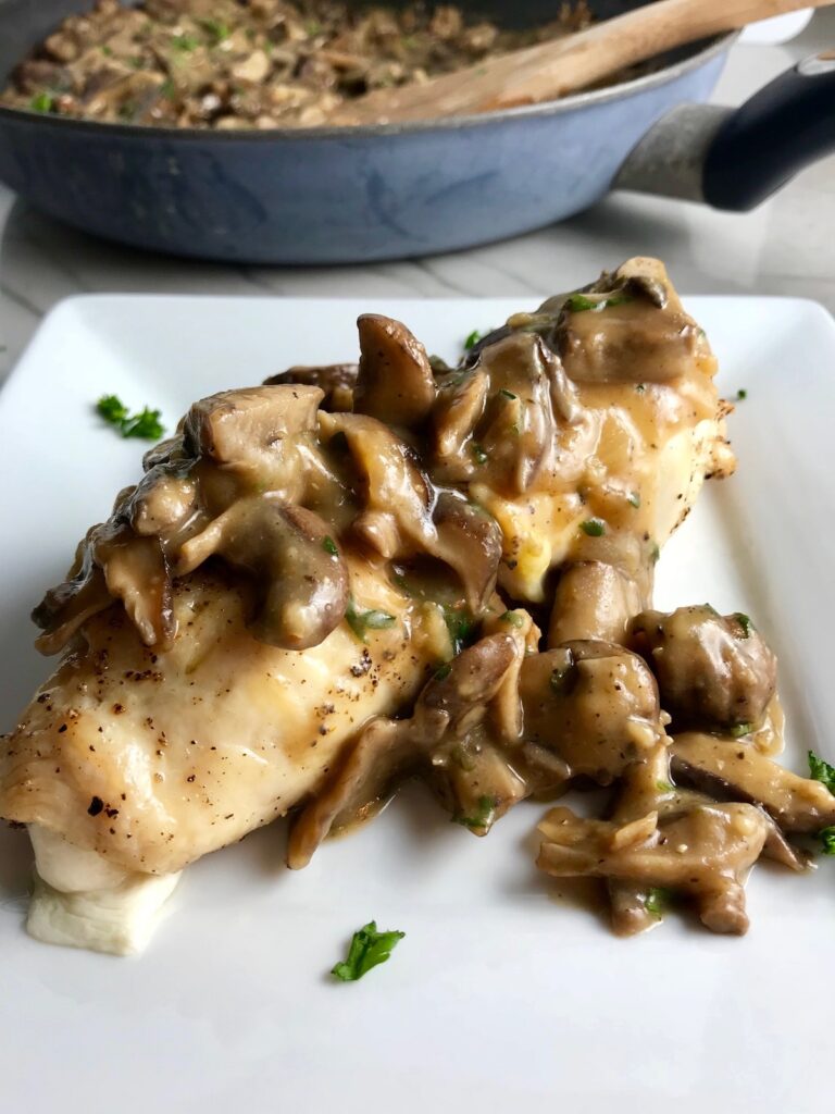 Mozzarella stuffed chicken roll on a plate with Mushroom sauce poured over top and parsley garnish sprinkled on top.  In the background is the skillet with the rest of the recipe and a wood spatula.