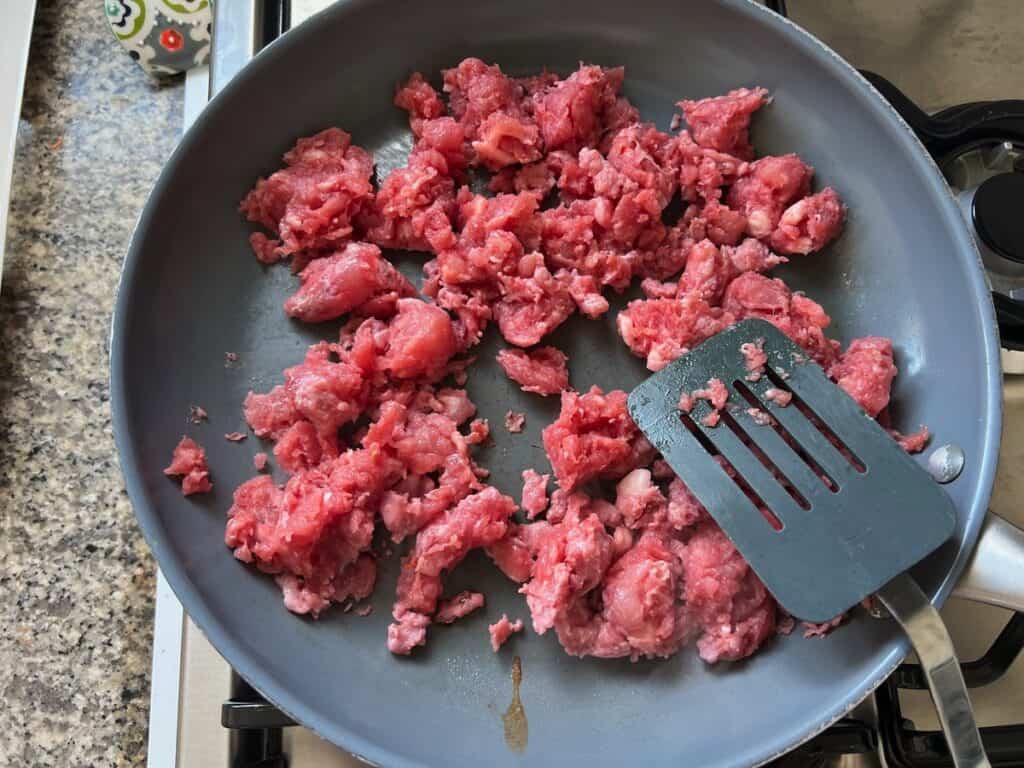 Spatula breaking up raw ground pork in a frying pan for Ground Pork Slow Cooker Casserole.