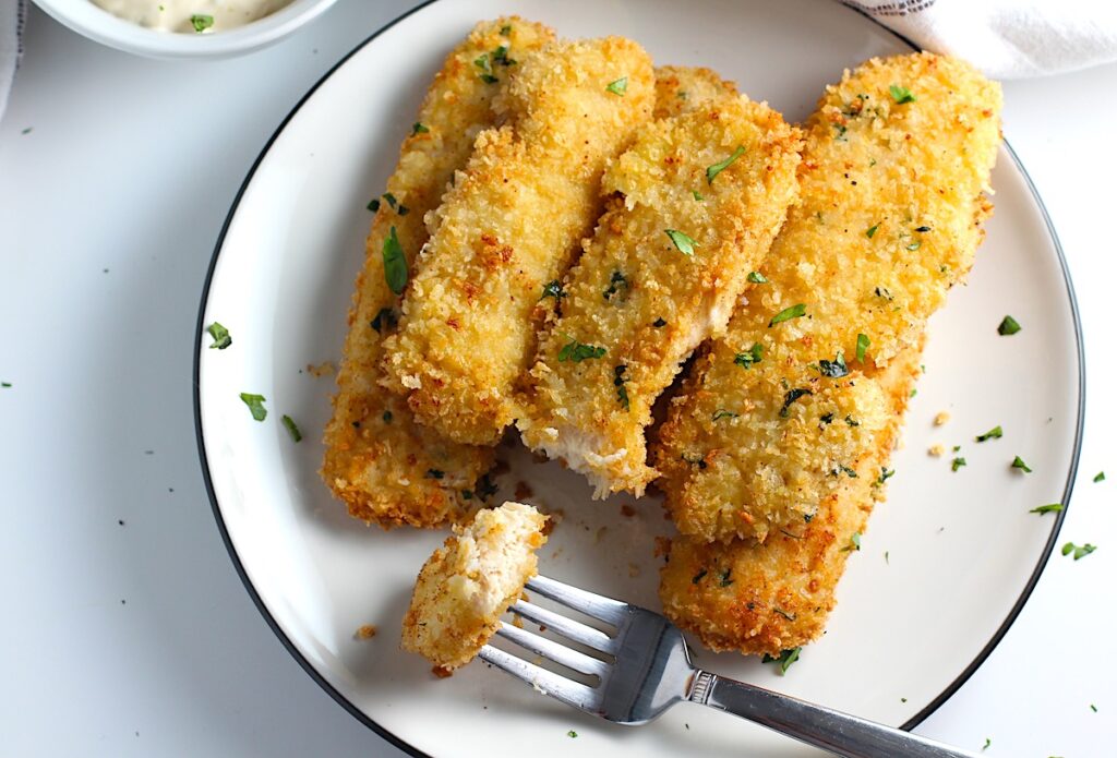 Several crispy and golden brown Panko Chicken strips stacked on a plate with a fork holding a bite.