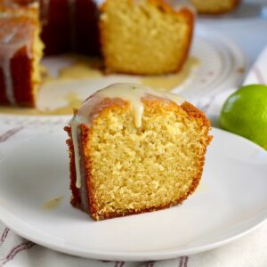Moist Vanilla Cake with Lime slice standing on a plate with lime glaze dripping down. A green lime behind the plate and the rest of the bundt cake on a platter in the background.