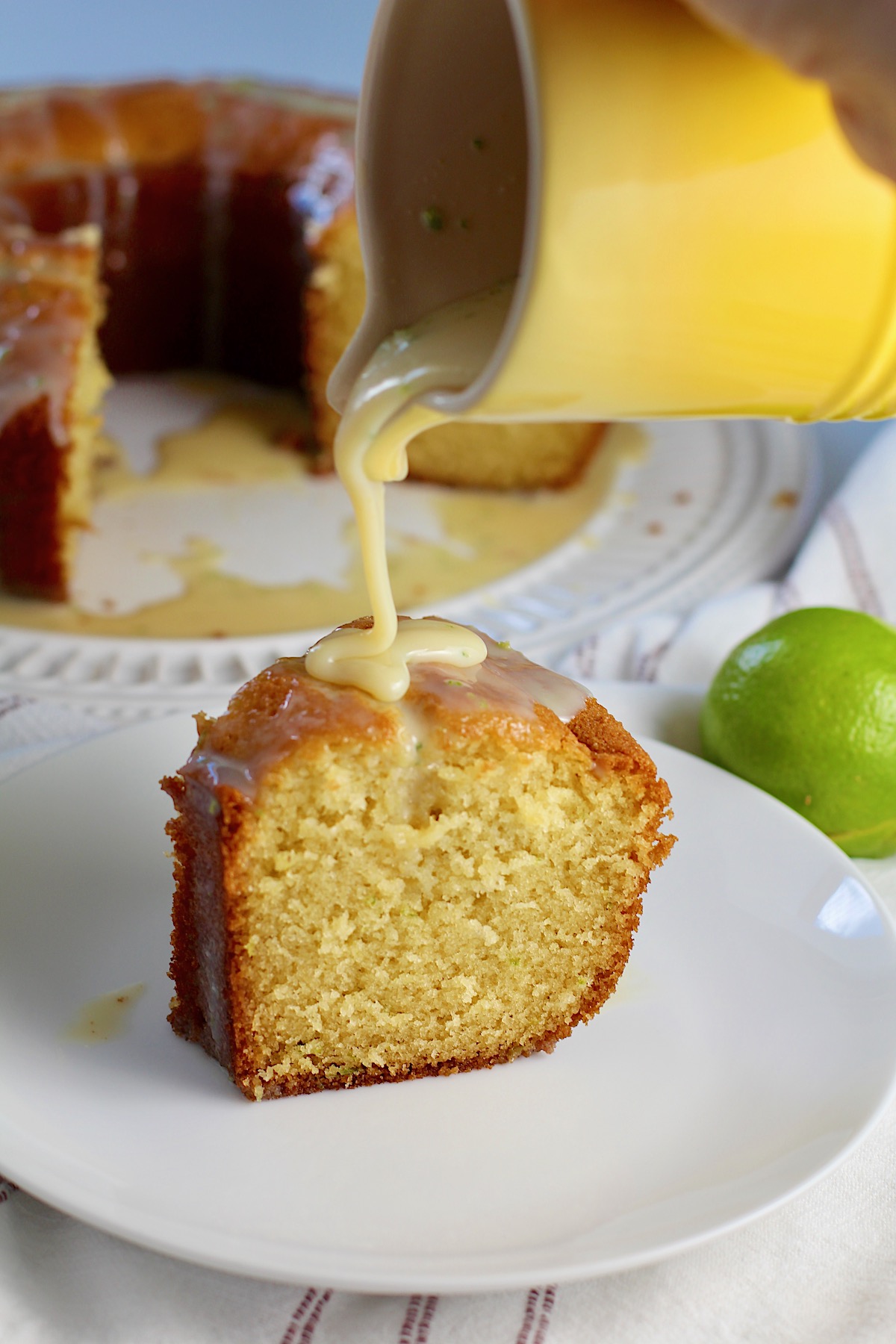 Yellow server pouring lime glaze over a slice of Vanilla Cake with Lime standing on a plate with lime glaze dripping down. A green lime behind the plate and the rest of the bundt cake on a platter in the background.