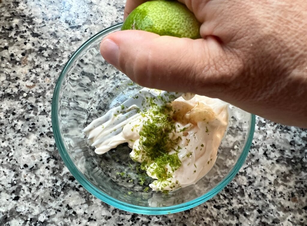 Hand squeezing lime into a bowl with mayonnaise, lime zest, and minced garlic for Lime aioli.
