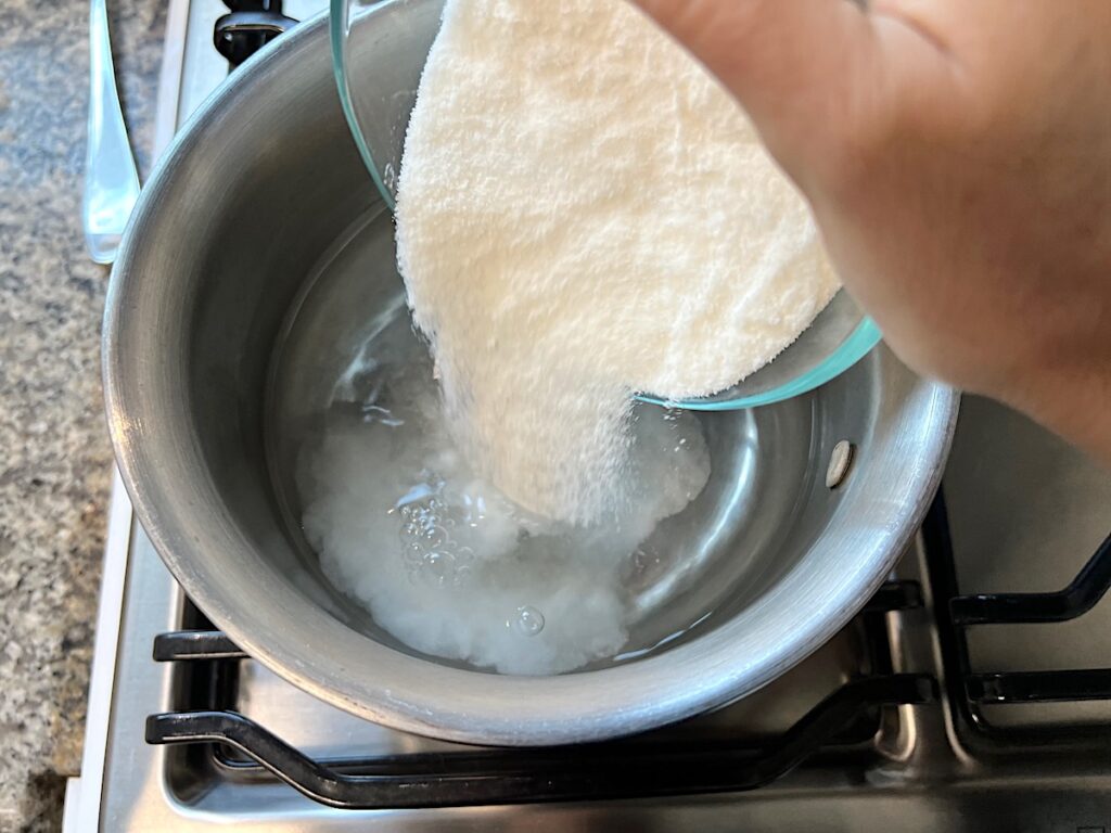 Hand pouring granulated sugar into water in a pot on the stove for simple syrup for How to make Strawberry Acai Refresher.